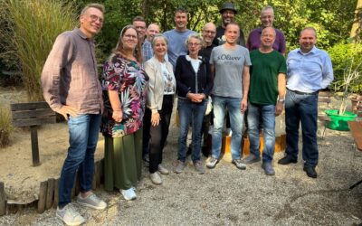 Zwölf lächelnde Männer und Frauen, ehemalige Klassenkameraden, stehen eng zusammen im Freien vor Grünpflanzen