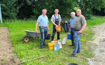 Vier ältere Männer mit Schubkarre und Eimern stehen auf einem umgegrabenen Feld neben einer Wiese und Bäumen