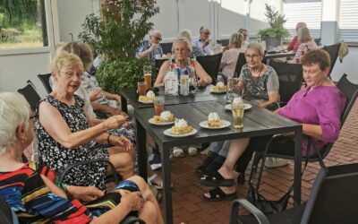 Eine Gruppe Senioren sitzt auf einer überdachten Terrasse an Tischen mit Kuchen und Getränken