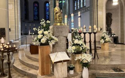 Altarraum einer Kirche mit goldener Marienstatue, weißen Blumen, brennenden Kerzen und einem geöffneten Buch