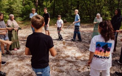Jugendliche halten im Wald bunte Seile, die zu einem Holzklotz in der Mitte führen, für ein Teamspiel
