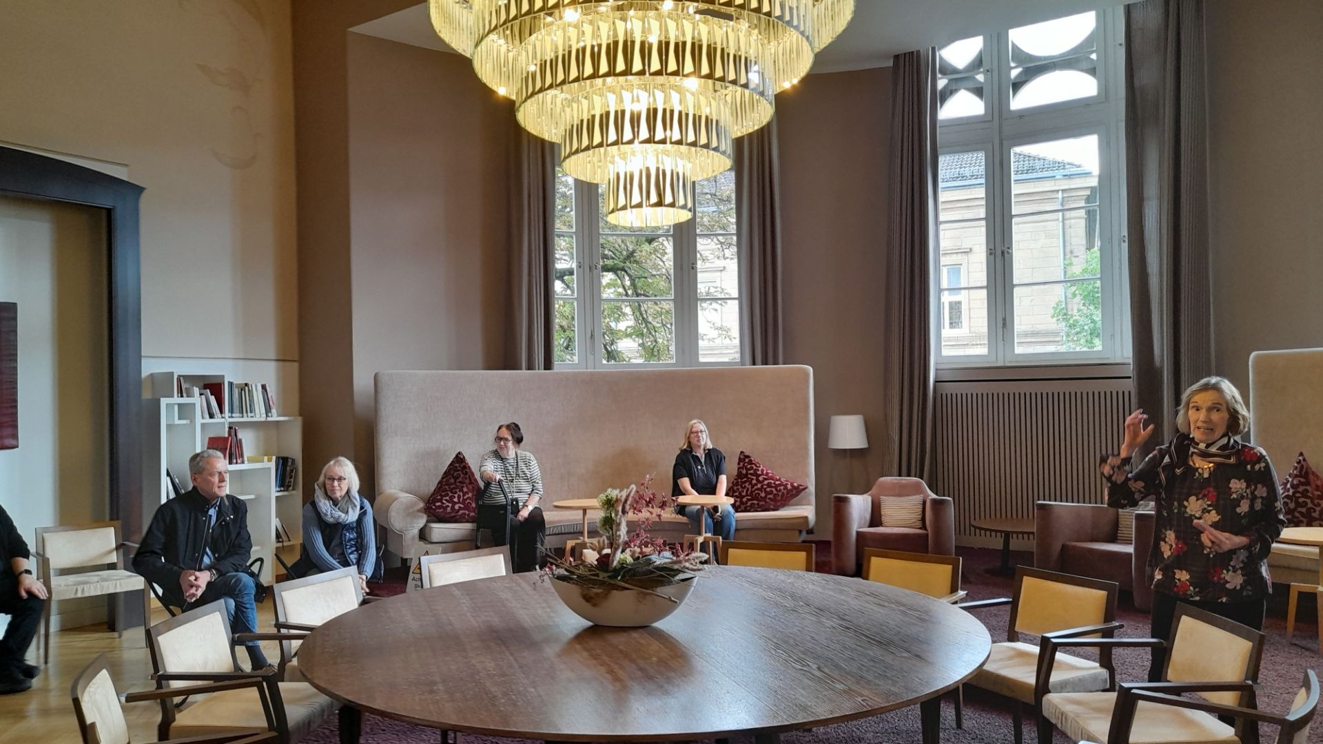 Eine Frau spricht gestikulierend vor sitzenden Personen in einem eleganten Raum mit Holztisch und großen Fenstern
