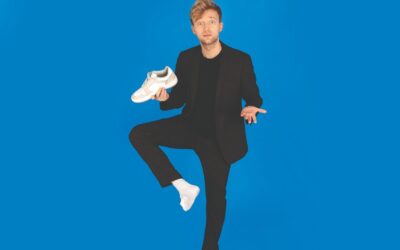 Marc Weide im schwarzen Anzug steht auf einem Bein, hält einen weißen Sneaker und schaut erstaunt vor blauem Hintergrund