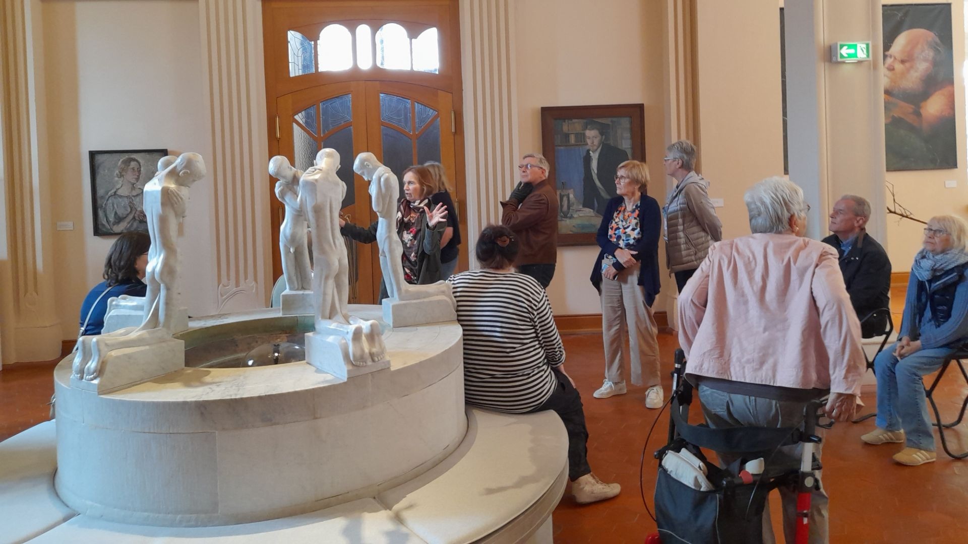 Ältere Besucher betrachten einen großen Marmorbrunnen mit menschlichen Skulpturen in einem Museumssaal mit Gemälden an