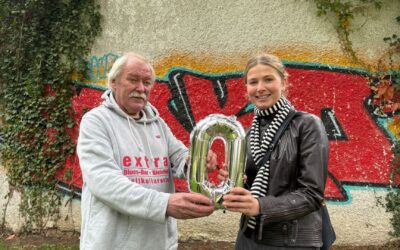 Ein Mann und eine Frau halten gemeinsam einen silbernen „0“-Folienballon vor einer bunten Graffitiwand in Bielefeld
