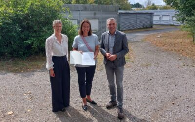 Zwei lächelnde Frauen und ein Mann stehen auf einem Kiesweg vor grauen Gebäuden, eine Frau hält ein Dokument
