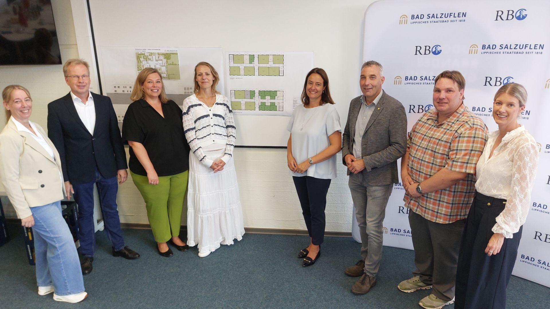 Acht Personen, vier Frauen und vier Männer, lächeln stehend vor Architekturplänen und einem Bad Salzuflen Logo