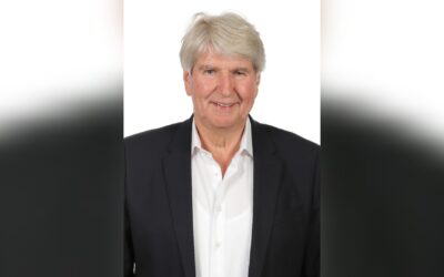 Ein lächelnder älterer Mann mit grauem Haar, dunklem Sakko und weißem Hemd vor weißem Hintergrund