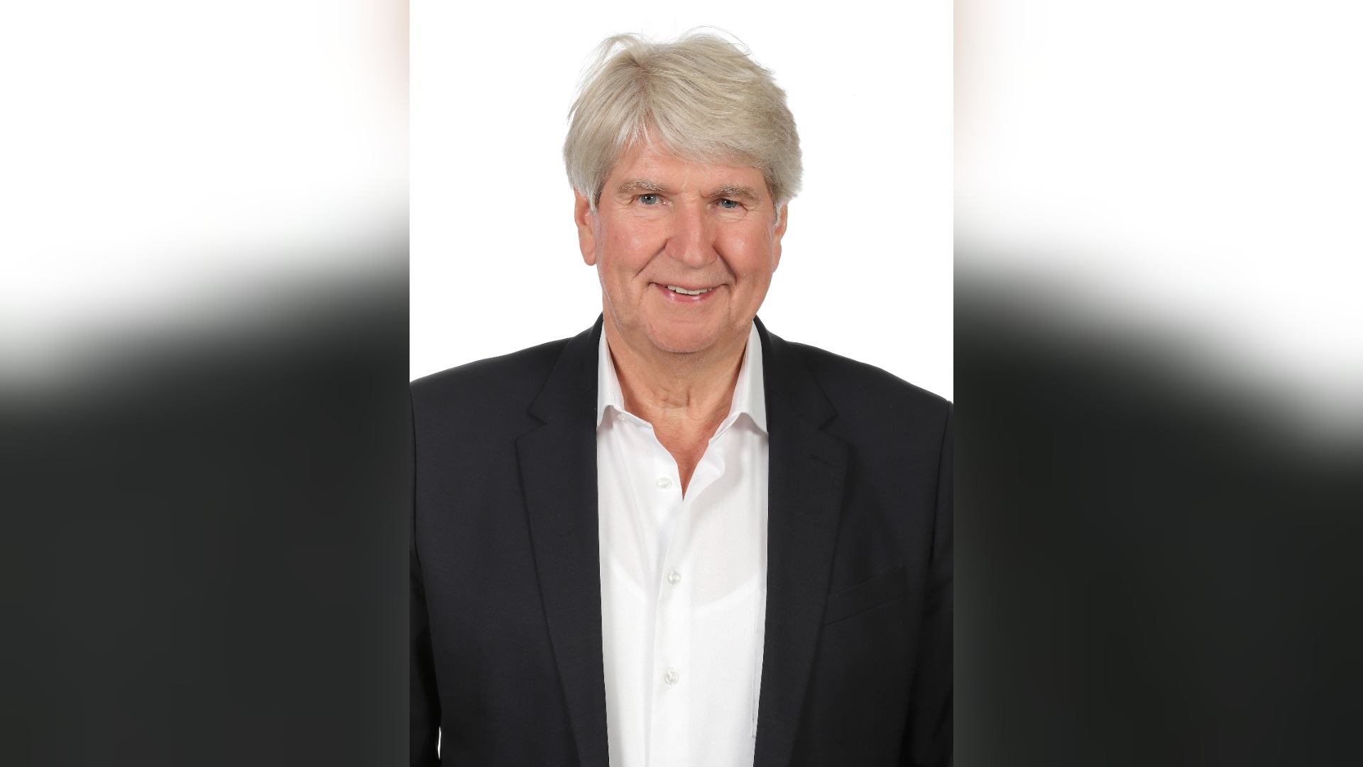 Ein lächelnder älterer Mann mit grauem Haar, dunklem Sakko und weißem Hemd vor weißem Hintergrund