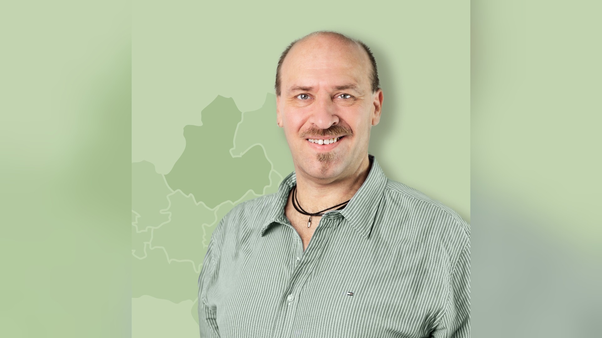Michael Böttcher, ein lächelnder Mann mit Schnurrbart und Glatze in gestreiftem Hemd vor einer grünen Karte