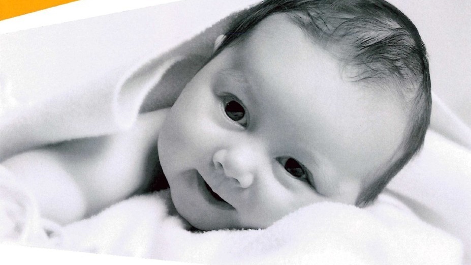 Kleines Baby mit dunklem Haar liegt auf einem weißen Tuch, lächelt leicht und schaut in die Kamera
