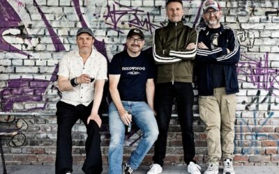 Vier Bandmitglieder der Seltaebs posieren vor einer graffitibesprühten Ziegelwand, zwei sitzen, zwei stehen mit