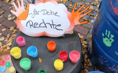 Schild "für Deine Rechte" mit orangenen Händen, davor bunte Farbtöpfe, daneben ein blaues Fass mit Handabdrücken