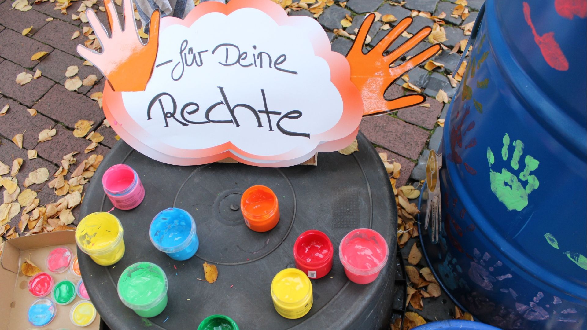 Schild "für Deine Rechte" mit orangenen Händen, davor bunte Farbtöpfe, daneben ein blaues Fass mit Handabdrücken