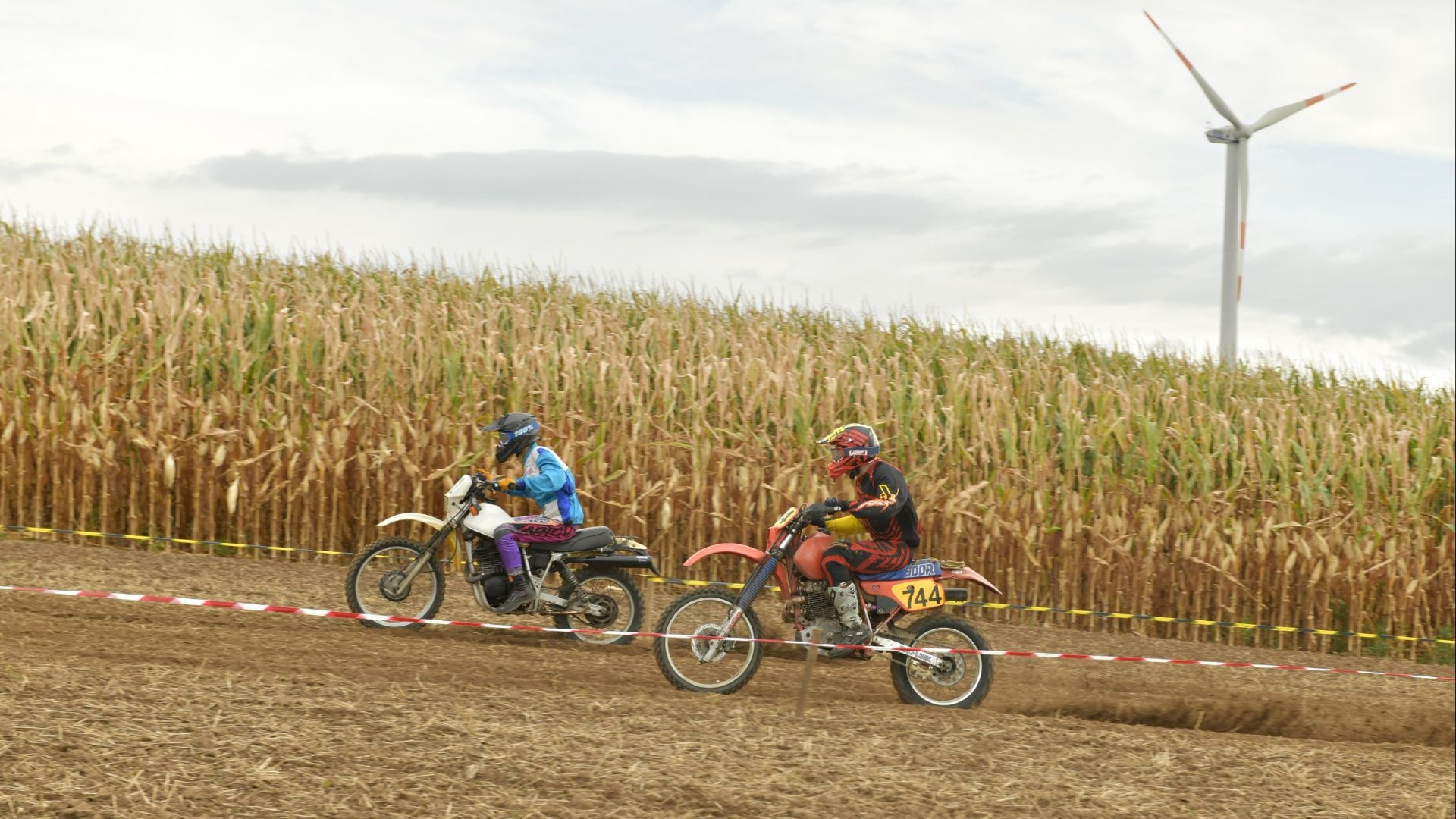 Zwei Enduro-Fahrer auf ihren Motorrädern fahren auf einer Erdstrecke neben einem Maisfeld mit Windrad