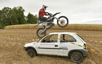 Motocrossfahrer springt mit seinem Motorrad im Wheelie über ein weißes Auto, das als Hindernis auf einer Geländestrecke