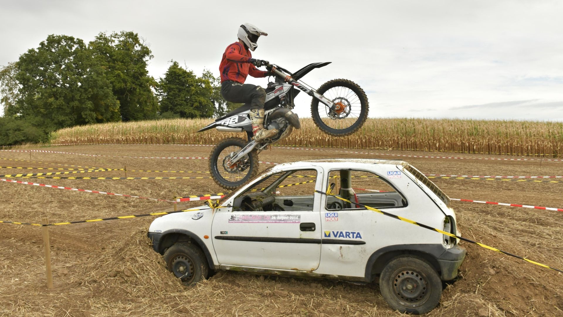 Motocrossfahrer springt mit seinem Motorrad im Wheelie über ein weißes Auto, das als Hindernis auf einer Geländestrecke