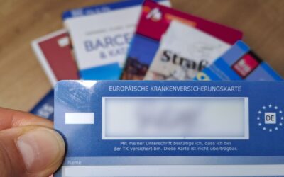 Eine Hand hält eine blaue Europäische Krankenversicherungskarte mit DE-Symbol, im Hintergrund unscharf weitere Karten