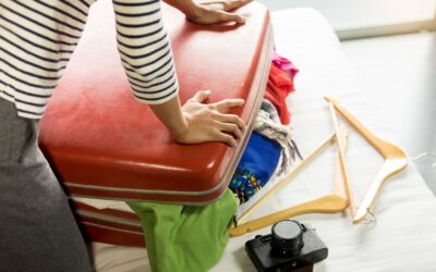 Person drückt auf roten, überfüllten Koffer auf einem Bett mit Kleidung, Kleiderbügeln und Kamera daneben