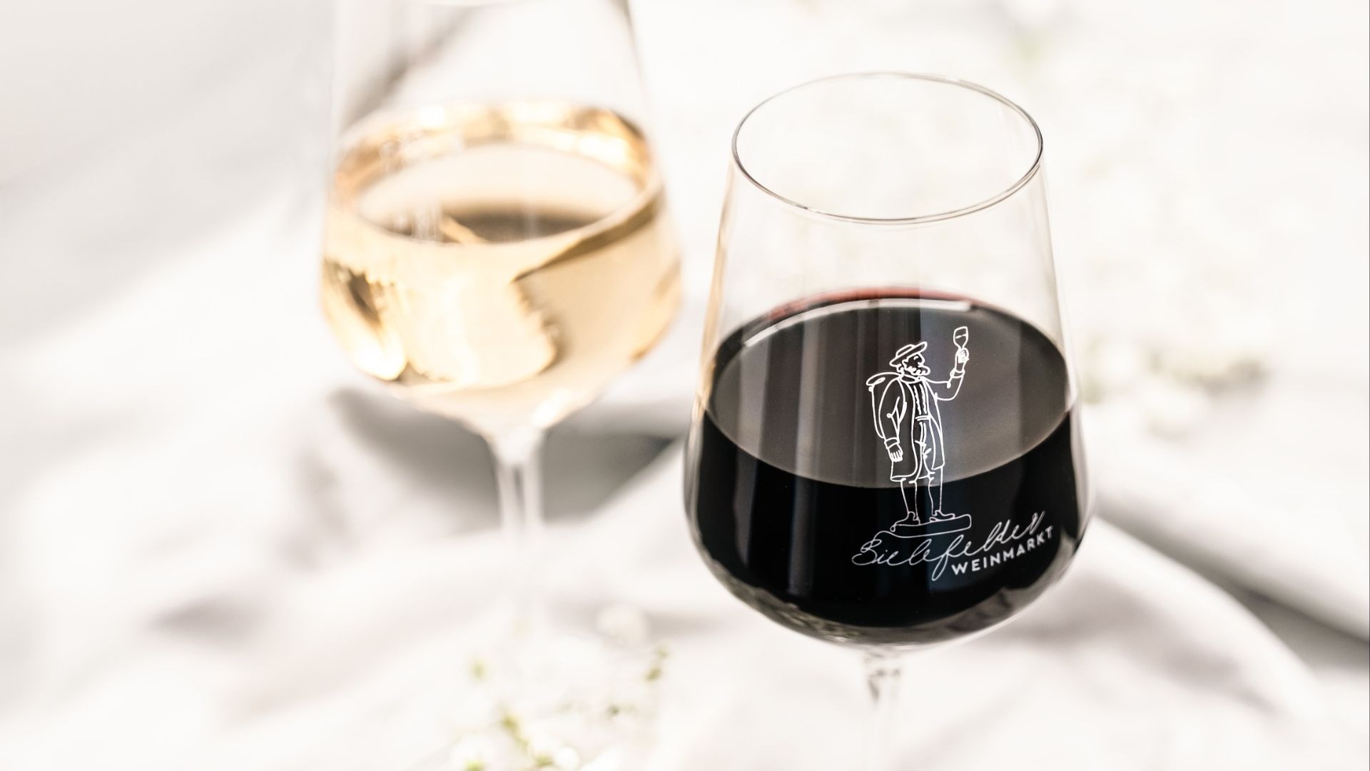 Zwei Weingläser mit Rot- und Weißwein, das vordere Rotweinglas trägt das Logo des Bielefelder Weinmarktes