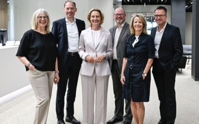 Sechs lächelnde Personen, drei Männer und drei Frauen, stehen in einem modernen Beratungszentrum