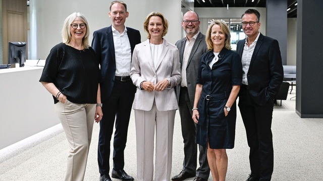 Sechs lächelnde Personen, drei Männer und drei Frauen, stehen in einem modernen Beratungszentrum