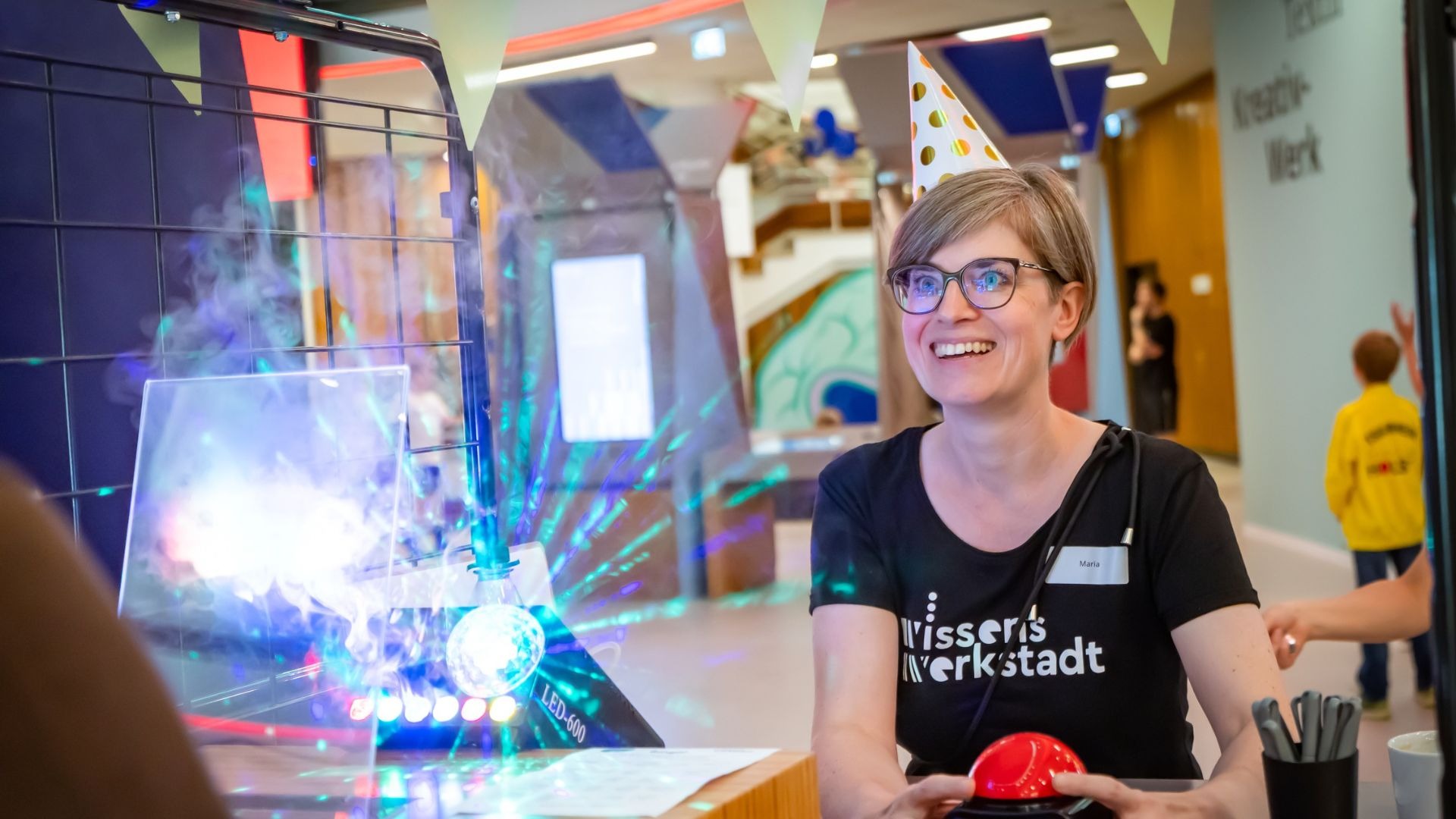 Lächelnde Frau Maria mit Partyhut und Wissenswerkstadt-Shirt sitzt an einem Tisch mit rotem Knopf, umgeben von bunten