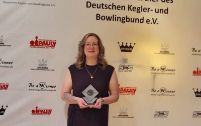 Christina Rähse mit Brille und dunklem Kleid steht vor einer Sponsorenwand und hält eine gläserne Auszeichnung