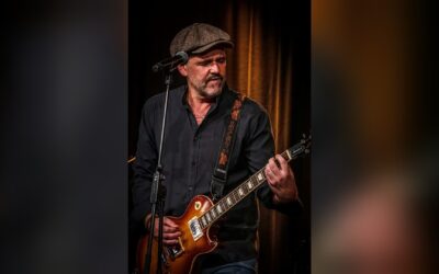 Mann mit Bart und Kappe spielt E-Gitarre an einem Mikrofon auf dunkler Bühne