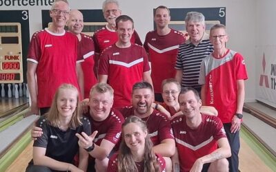 Eine Gruppe lachender Sportkegler in roten Trikots posiert auf einer Kegelbahn mit Pins und Anzeigetafel