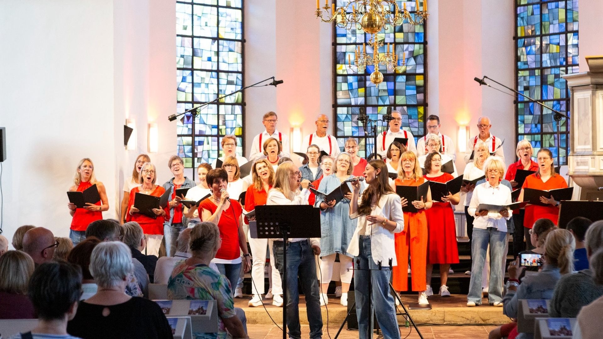 Ein großer gemischter Chor singt mit Notenheften in einer hellen Kirche, zwei Personen am Mikrofon führen den Gesang,