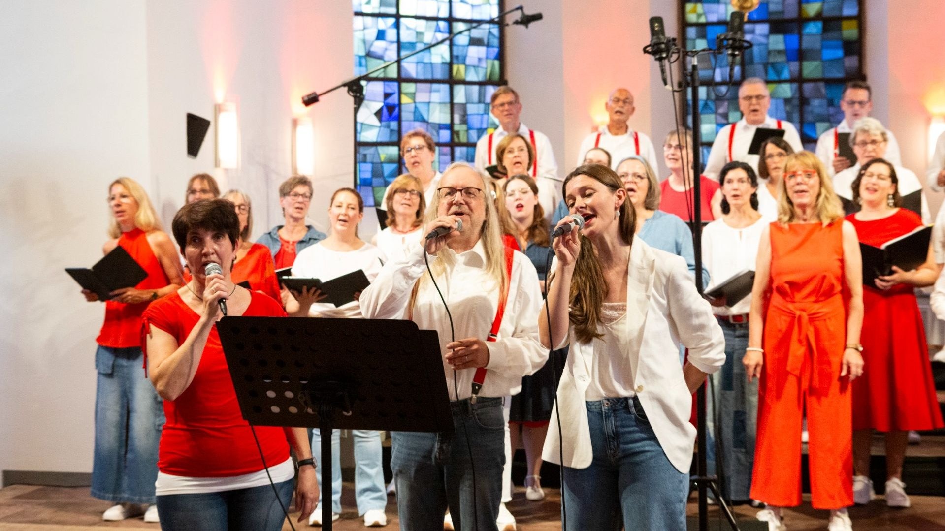 Mehrere Erwachsene singen in einem Chor in einer Kirche, einige mit Mikrofonen vor einem Buntglasfenster