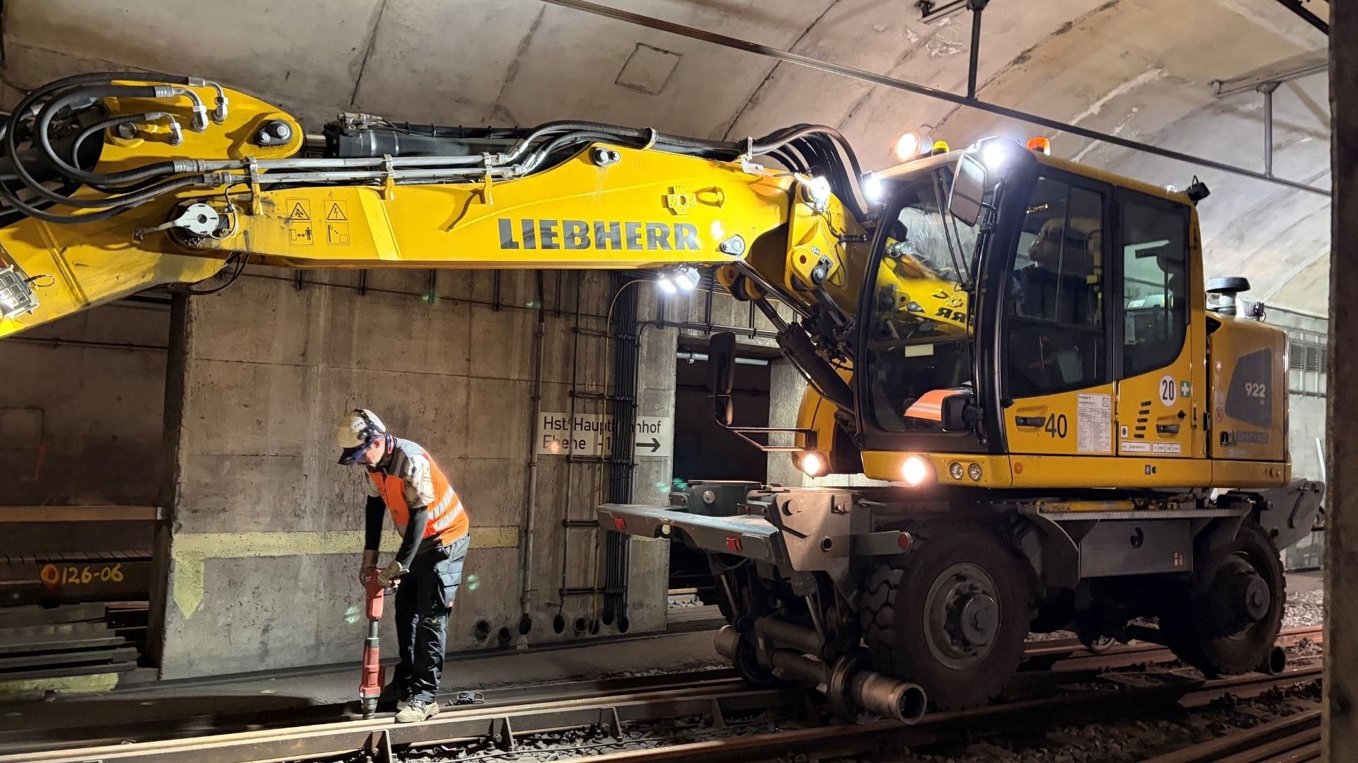 Ein Arbeiter in oranger Warnweste bedient einen Bohrhammer neben einem gelben Schienenbagger in einem Stadtbahntunnel