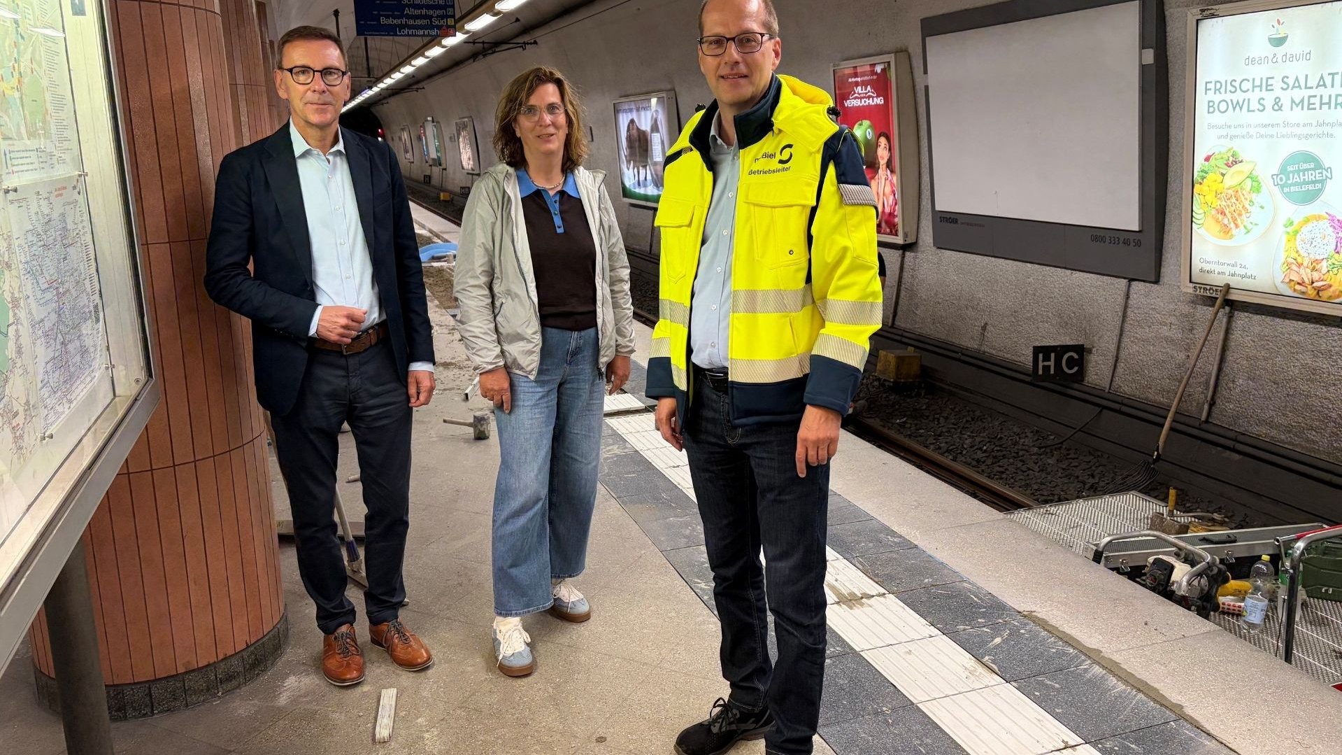 Drei Personen, ein Mann im Anzug, eine Frau in Jeans und ein Mann in gelber Warnweste, auf einem Bahnsteig im