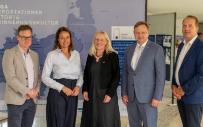 Vier Männer und eine Frau stehen lächelnd vor einer Ausstellung zu Deportationen und dem Riga-Komitee