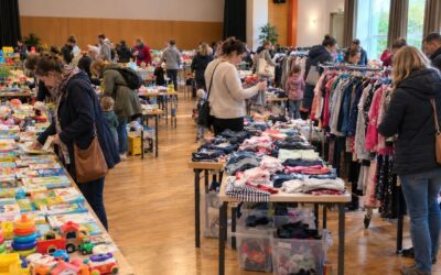 Zahlreiche Erwachsene und Kinder stöbern an Tischen voller Kleidung, Spielzeug und Büchern auf einem Kinderflohmarkt in