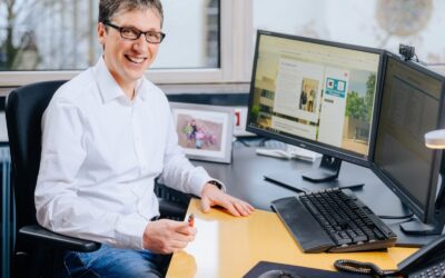 Lächelnder Mann mit Brille und weißem Hemd sitzt an einem Schreibtisch mit zwei Monitoren, Telefon und Familienfoto