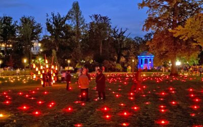 Dunkler Park mit Menschen, die rote Bodenlichter, eine hohe Laternenkonstruktion und einen blau leuchtenden Pavillon