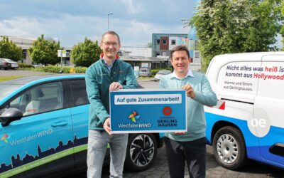 Zwei lächelnde Männer halten ein Schild "Auf gute Zusammenarbeit" zwischen einem WestfalenWIND Auto und einem