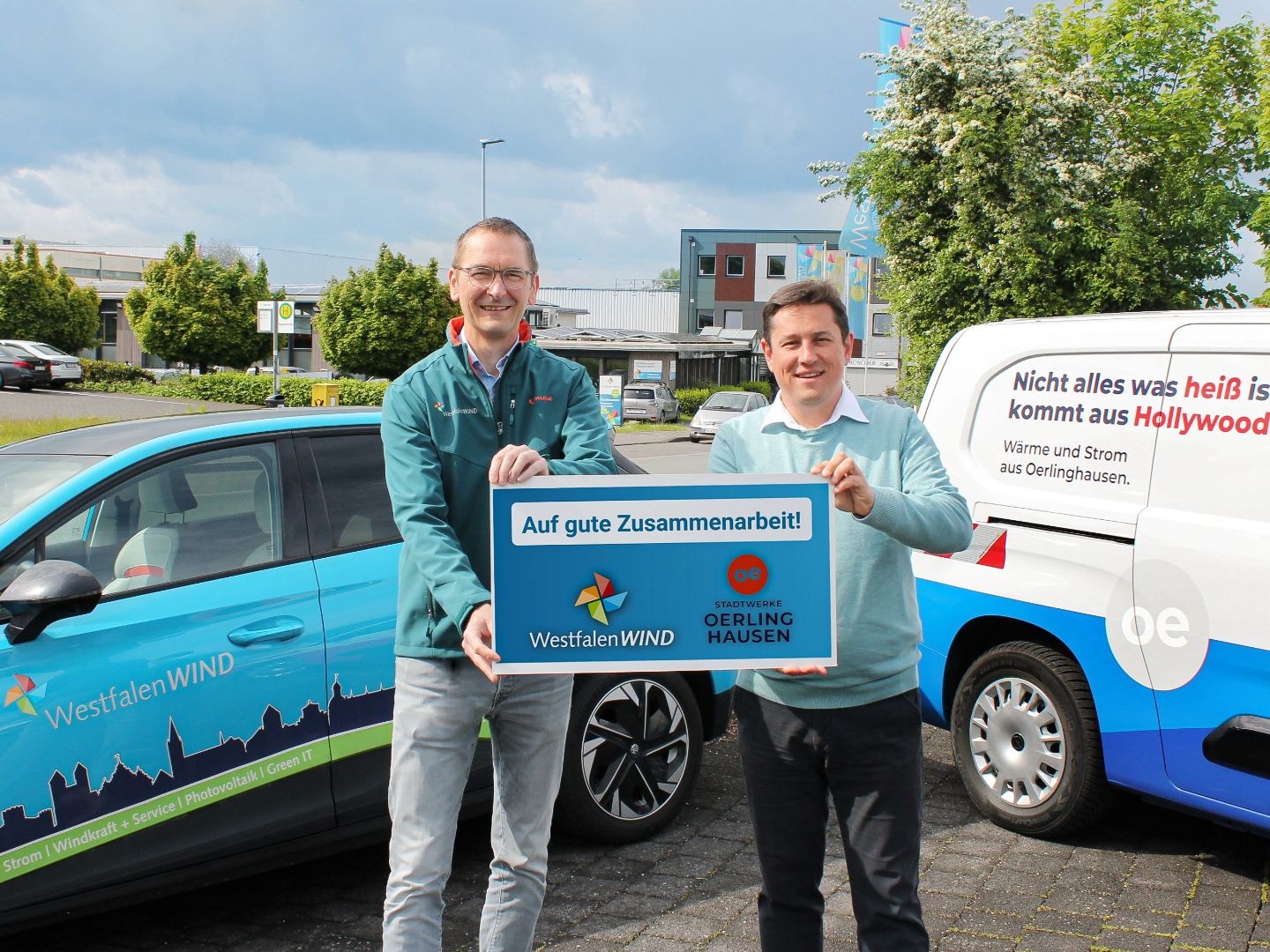 Zwei lächelnde Männer halten ein Schild "Auf gute Zusammenarbeit" zwischen einem WestfalenWIND Auto und einem