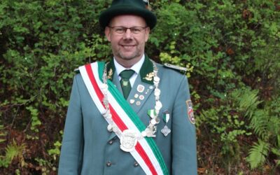 Mann in grüner Schützenuniform mit Orden und Schützenkette lächelt vor Grünpflanzen