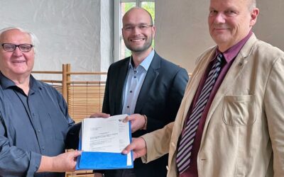 Drei lächelnde Männer überreichen einen Förderbescheid in einem Besprechungsraum