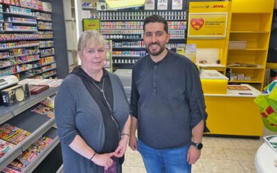 Eine blonde Frau und ein bärtiger Mann stehen lächelnd in einem Lotto- und Postladen vor Regalen