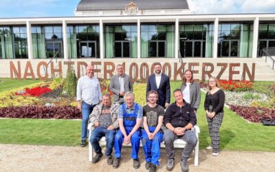 9 Personen stehen und sitzen vor einem Gebäude und einem Blumenbeet mit dem Schriftzug "Nacht der 1.000 Kerzen
