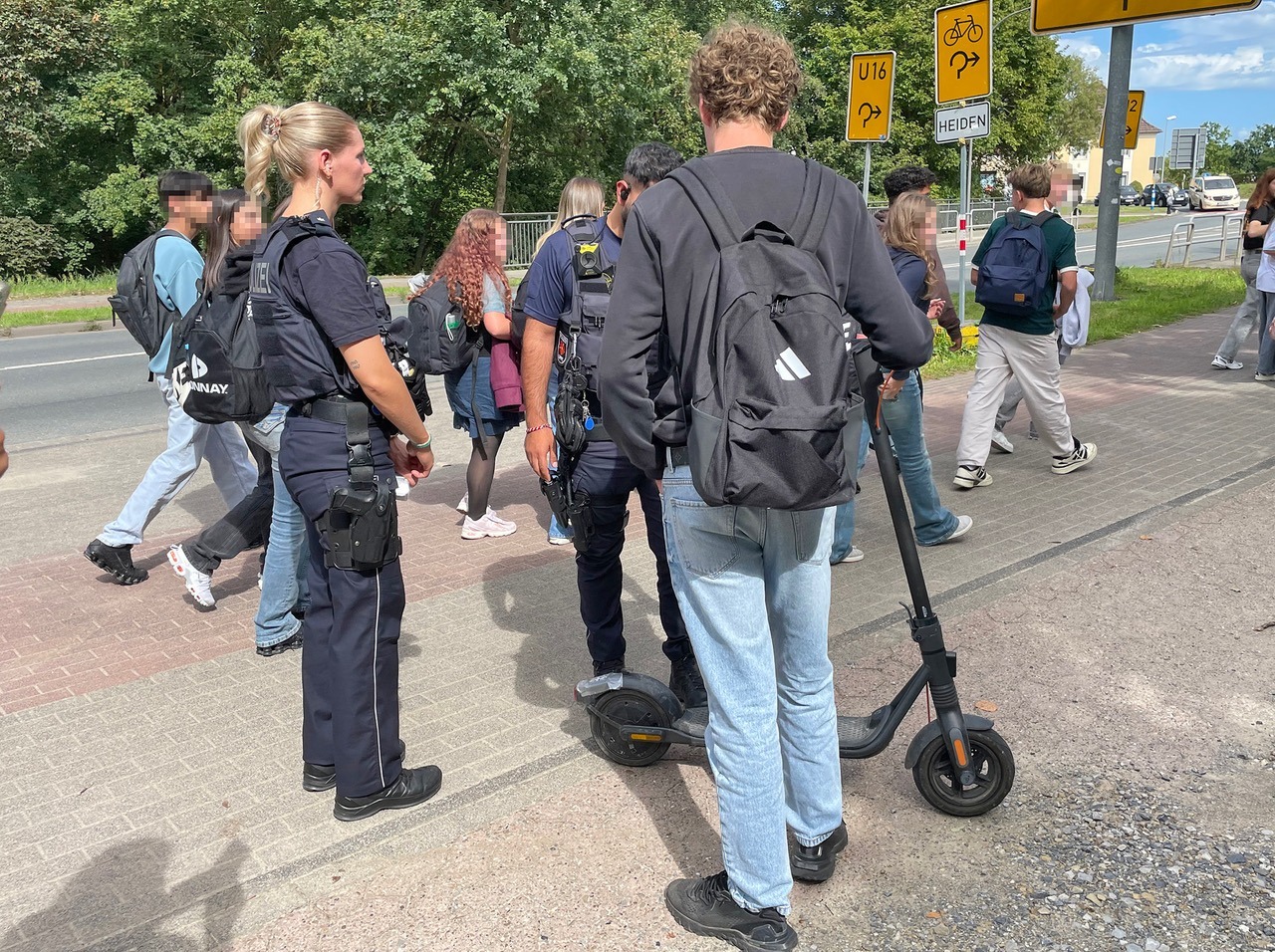 Polizisten kontrollieren mehrere Jugendliche und einen E-Scooter auf einem Bürgersteig neben einer Straße