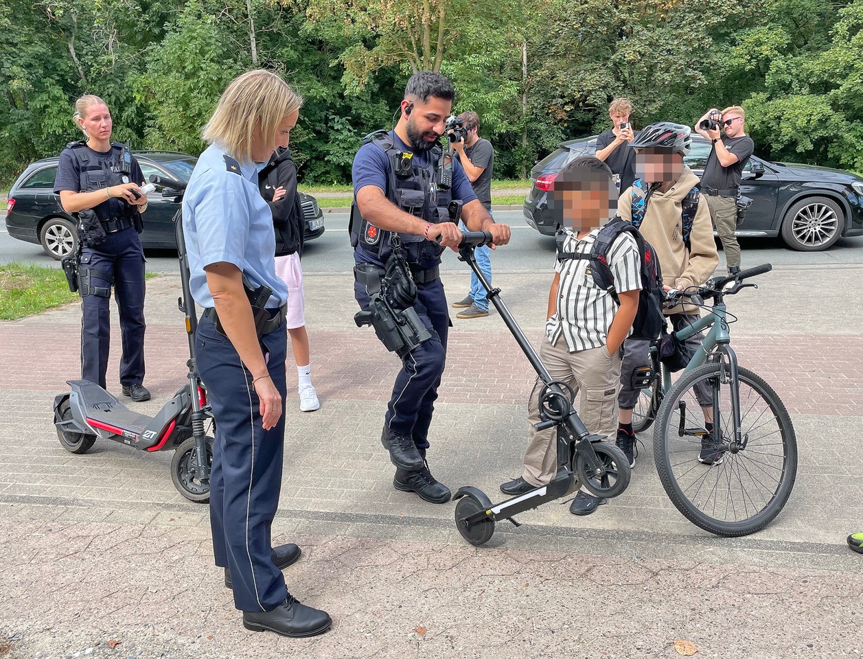 Polizisten kontrollieren zwei Jungen mit E-Scootern und einem Fahrrad am Straßenrand
