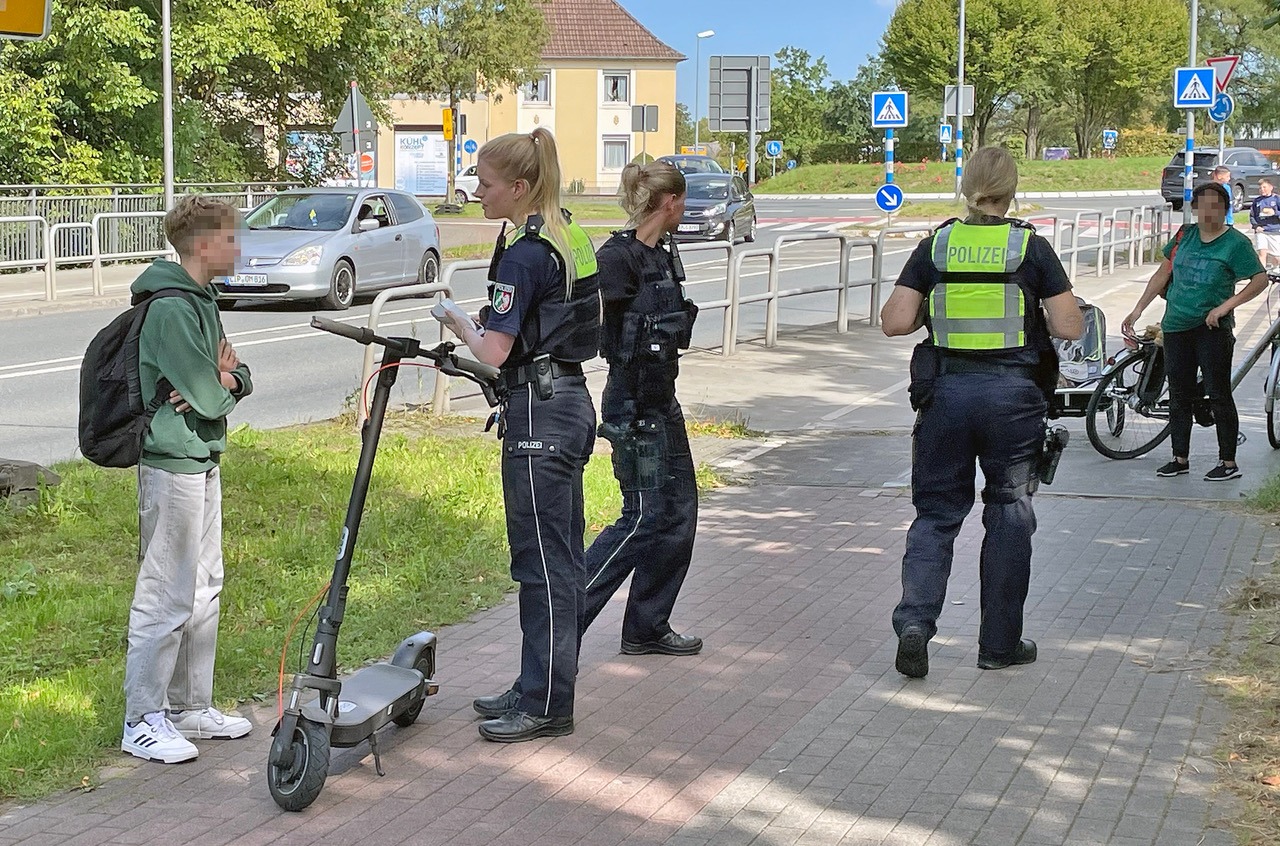 Drei Polizeibeamte kontrollieren Jugendliche mit E-Scooter und Fahrrad auf einem Gehweg neben einer Straße
