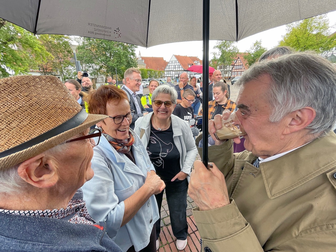 Innenminister Herbert Reul trinkt Kaffee unter einem Schirm und spricht lächelnd mit Bürgern auf dem Salzhof