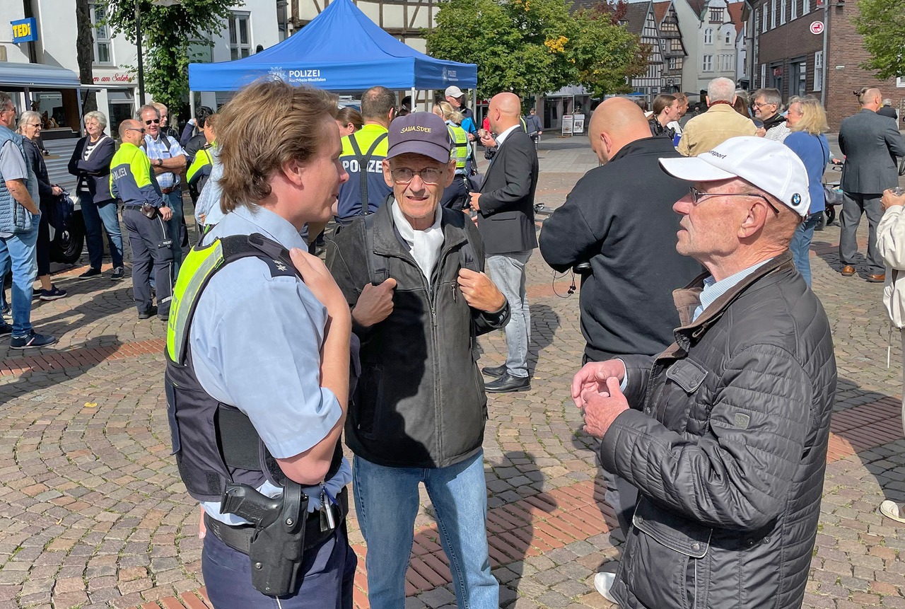 Ein Polizist unterhält sich auf einem belebten Platz mit zwei älteren Männern, ein Polizeizelt steht im Hintergrund