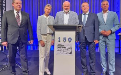 Vier Männer und eine Frau stehen auf einer Bühne vor einem Rednerpult mit einem 150-Jubiläums-Logo